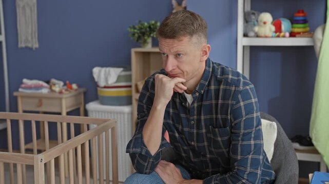 Man sitting with hand on chin next to wooden crib in nursery room inside building, gazing at empty cradle; quiet concern.