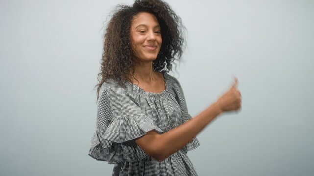 Teenage woman with curly hair points finger to right, smiling and showing forearm in studio wearing a checkered ruffled dress; playful confidence.