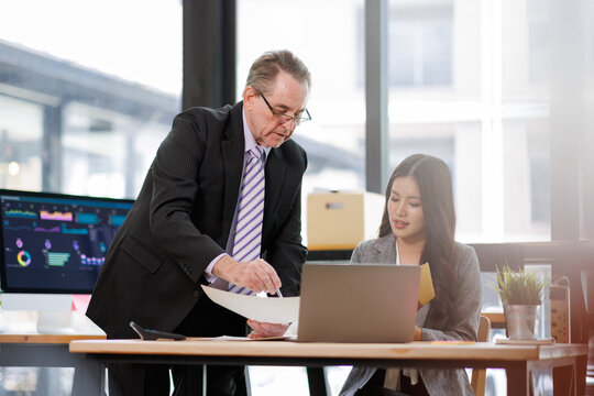 Business mentor and intern reviewing documents and data on a laptop, collaborating in a modern corporate office to coach skills, plan strategy, and support professional growth