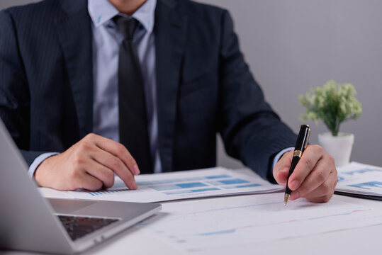Businessman hand holding pen analyzing financial document report with laptop at desk