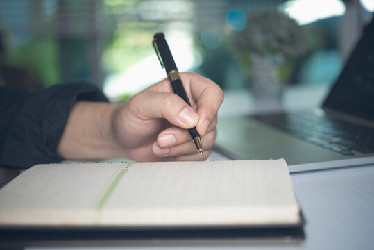 Hand holding pen writing in notebook with laptop on desk for business planning