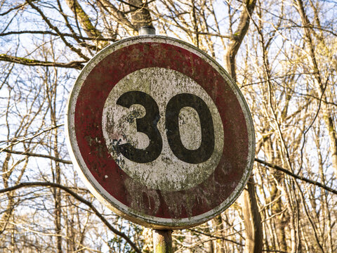 Old 30 kph speed limit sign, panneau de limitation de vitesse on a road in France, showing a weathered circular traffic sign surrounded by trees and highlighting driving rules and road safety.