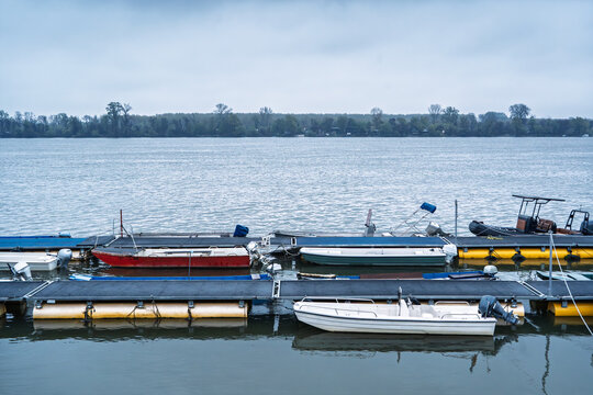Boats moored along the Danube near Zemunski kej in Belgrade, Serbia, with floating docks, calm river water, cloudy weather, and the opposite riverbank visible in the distance.