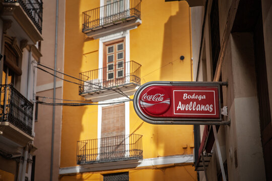 VALENCIA, SPAIN - APRIL 6, 2025: Coca Cola sign mounted outside Bodega Avellanas in Valencia, Spain, with a red beverage logo sign set against a yellow building facade and old town street.