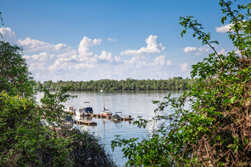 Boats anchored along Zemunski kej on the Danube River in Zemun, Belgrade, Serbia, with a small dock, calm water, green riverbank, and blue sky. © Jerome