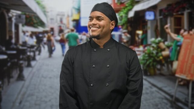 Man in chef uniform with smiling face looks to side on a cobblestone street outside a cafe; pride hospitality.