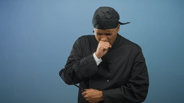 Hispanic man chef with beard in black uniform coughing into fist while holding spatula in studio; fatigue.