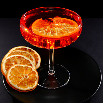 Vibrant red cocktail with ice cubes in a coupe glass, accompanied by dried orange slices, captured in a striking studio photograph with dramatic lighting against a dark background closeup view 
