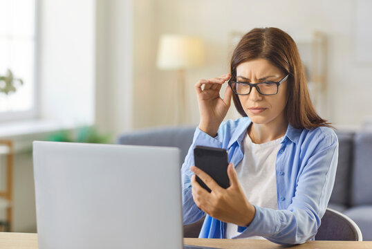 Woman with glasses squinting at smartphone screen, sitting at home office desk with laptop. Person with poor eyesight squinting through eyeglasses to struggle with eyestrain, check blurry text