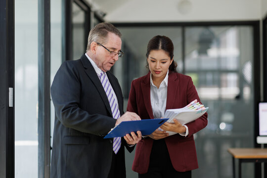 Mature business professional mentoring asian female junior executive, discussing paperwork and documents, collaborating on a project in a modern corporate office environment
