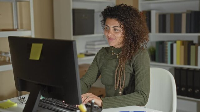 Woman at computer with hand on cheek, smiling and typing at desk in building with bookshelves and monitor visible; contentment productivity.
