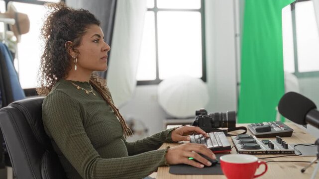 Woman typing on keyboard and using mouse while reaching to audio mixer and dslr camera in studio; focused creativity.