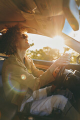 Fototapeta premium Smiling woman with curly hair driving a car during golden hour. Sunlight fills the cabin with a warm glow, creating a relaxed lifestyle moment focused on travel and self care.