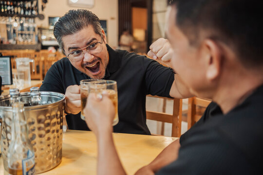 A lively and rowdy middle aged man enthusiastically toasts beer with his more reserved friend at a cozy, warm-lit bar, celebrating with joy.