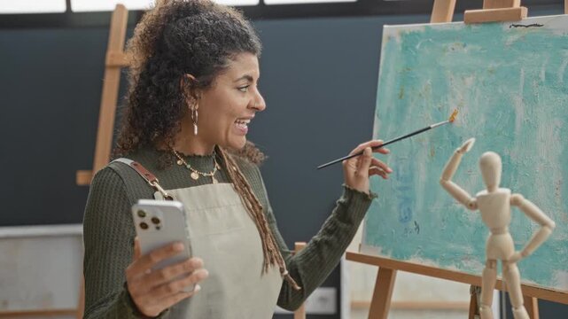 Woman painter in studio holds smartphone in left hand and paintbrush in right hand while pointing at canvas; creative focus.