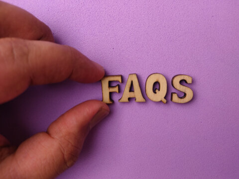 A hand holds the wooden word FAQS over a purple background surface