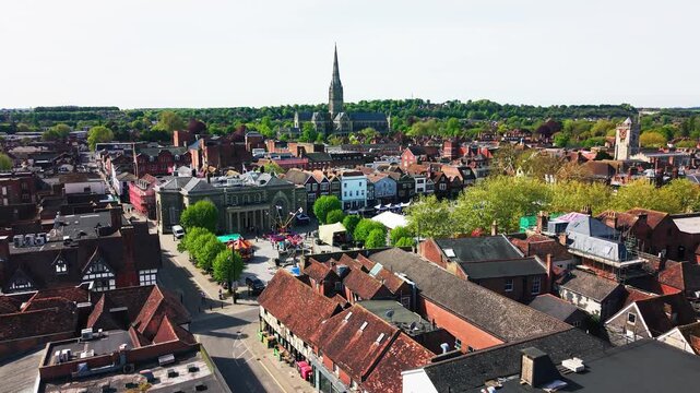 Aerial drone footage of Salisbury city centre with Salisbury Cathedral visible in the background, showing rooftops, historic streets, the Guildhall, Market Square, trees, traffic,