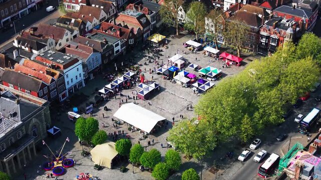 Aerial drone footage looking down over Salisbury Market Square during a St George&rsquo;s Day event, showing market stalls, crowds, fairground rides, trees, traffic, the Guildhall, historic buildings,