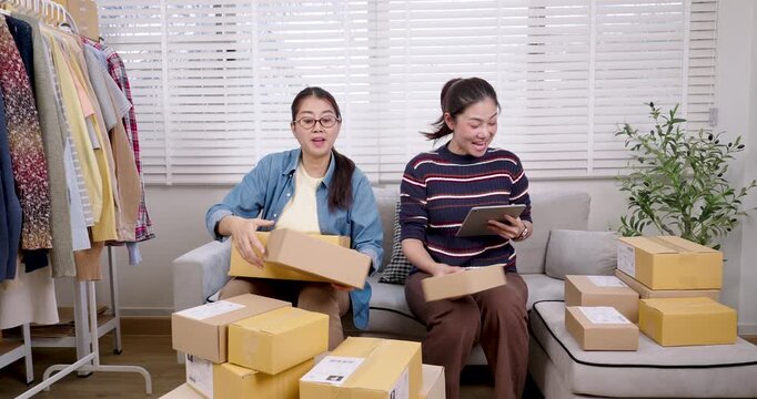 Asian women startup business partners lifting parcel box, placing packages on floor, checking online shopping parcel box list together, coordinating e-commerce order packing process inside home