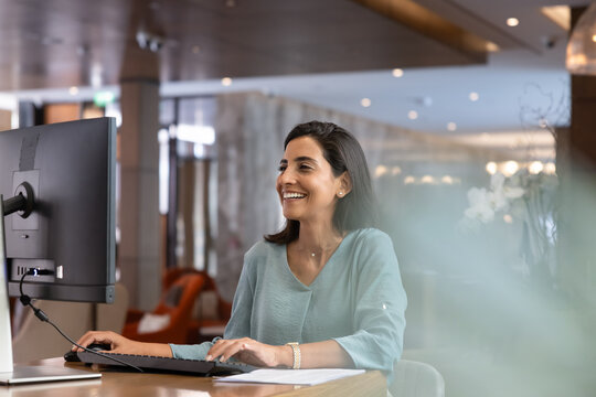 Smiling female receptionist working on computer at reception desk, verifying reservation details and guest records, preparing report, organizing hotel daily administrative workflow tasks at front desk