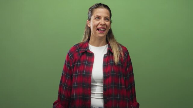 Woman in red plaid shirt smiling with teeth, head tilt and relaxed shoulders against green backdrop in studio; warm friendly joy.
