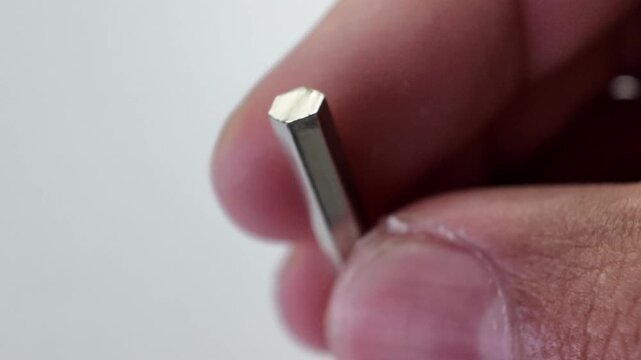 Close up view of a person's fingers reaching down to pick up a silver phillips cross head screwdriver bit from a collection scattered on a gray paper surface.
