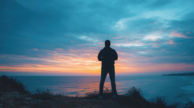 Solitude at Sunset: A lone figure stands silhouetted against a vibrant sunset, gazing out at the vast expanse of the ocean, embodying a moment of contemplation and peace.