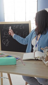 Woman writing letters on blackboard and pointing with chalk in classroom, seated at desk with notebook and colored pencils; focused learning.