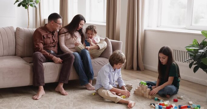 Siblings building with blocks on floor, parents and sister sitting on sofa using smartphone, large family spending leisure, communicating, play games, relaxing together at home. Childhood development