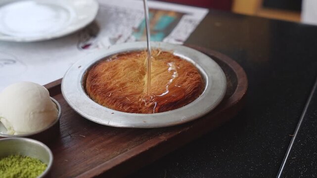 Close-up of clear sugar syrup being poured from a copper pot over a warm Middle Eastern Kunafa. Traditional knafeh served with vanilla ice cream and pistachios on a wooden tray.