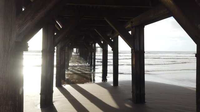 Handheld View Under Wooden Pier At Beach With Sun Reflection And Ocean Waves