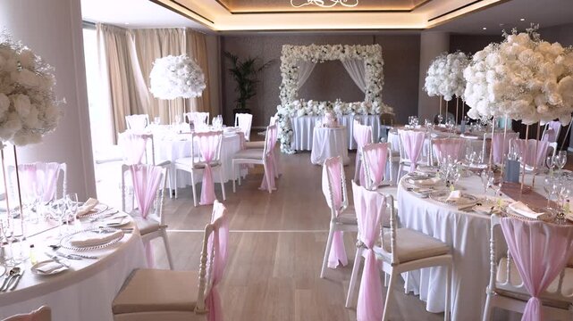 People gather in a banquet hall decorated with floral arrangements. Tables are set with white tablecloths and pink accents. The atmosphere shows preparations for a celebratory event.