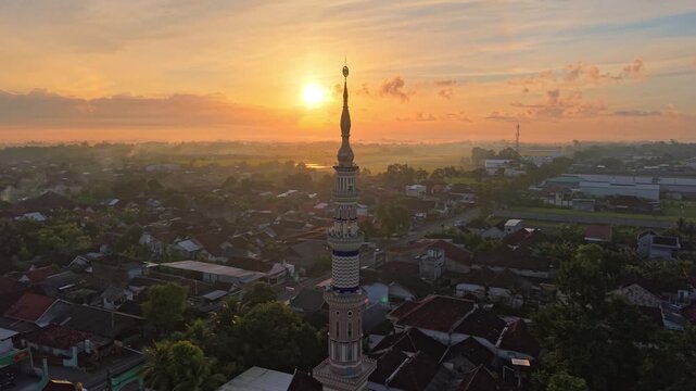 Drone shot of mosque tower against sunset sky, Islamic landmark, warm evening light, urban Indonesia scene.