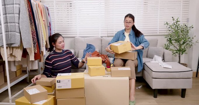 Asian women small business partners sitting with stacked parcel box, discussing order quantity, coordinating online shopping parcel box preparation together inside home based e-commerce warehouse