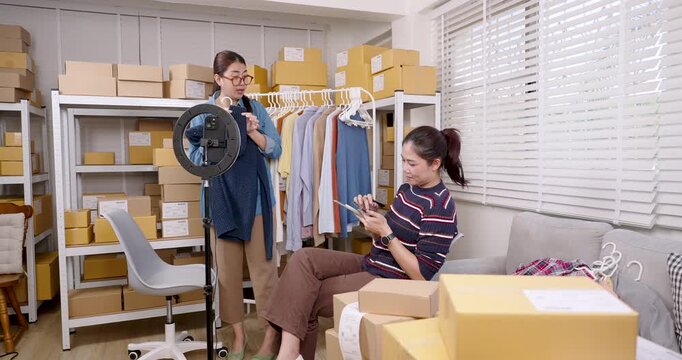 Asian women online business partners adjusting clothing display, preparing apparel for live selling, reviewing product detail on tablet inside home based e-commerce warehouse studio
