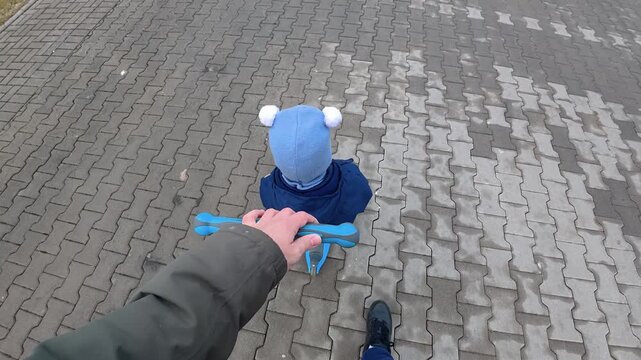 A person guides a child riding a blue tricycle on a wet cobblestone path. The overhead view captures a serene moment of bonding, with muted winter tones and soft lighting.