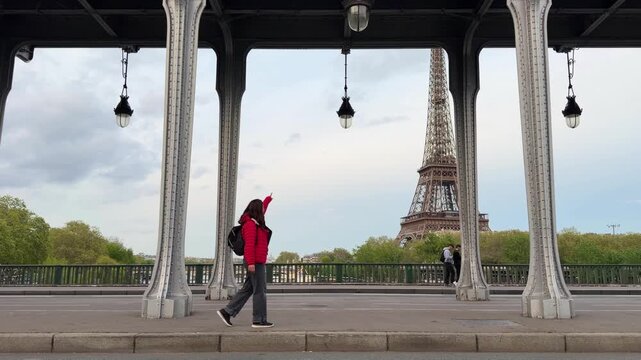 Young tourist woman in a red jacket walking along the famous Pont de Bir-Hakeim in Paris, pointing happily towards the iconic Eiffel Tower on a cloudy day in the french capital city