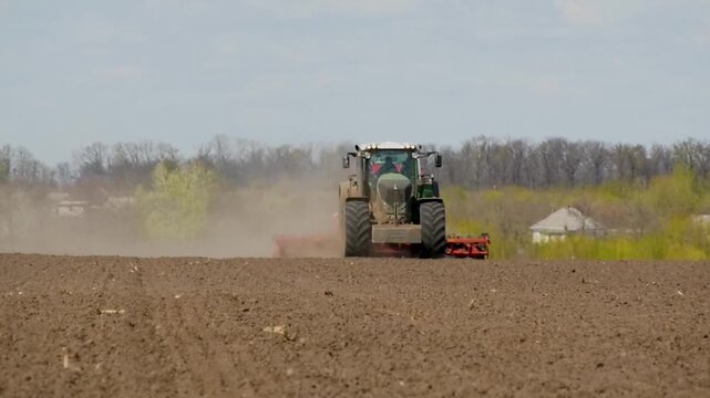Tractor driving through a field with a harrow and seeder, sowing wheat during spring field work. Dust rises behind the machinery during the sowing campaign. Concept of modern agriculture, grain