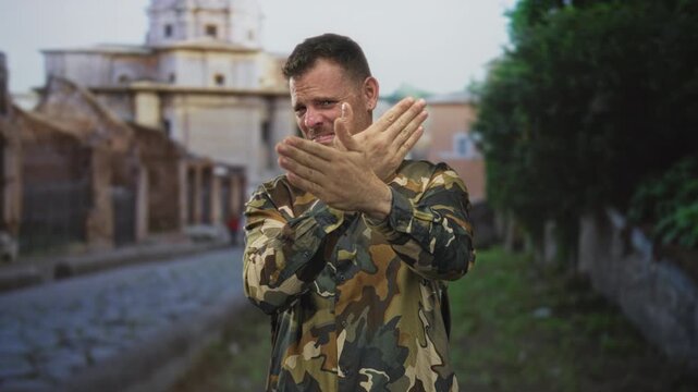 Man in camouflage uniform covers his face with both hands on a cobblestone street by an old building; grief reflection.