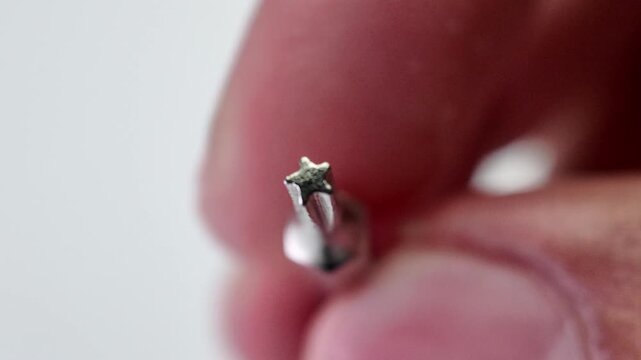 Close up view of a hand reaching down to select and pick up a long silver phillips cross head screwdriver bit from a neutral gray studio workbench backdrop.