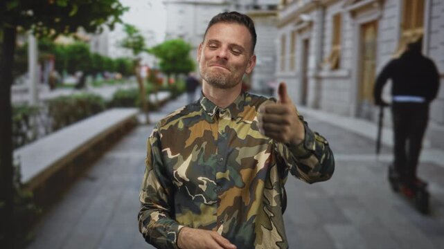 Man soldier in camouflage shirt giving thumbs up and smiling on a city street with bench and stone building; pride duty.