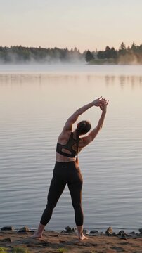 Woman stretching by misty lake at sunrise in yoga pose, vertical footage