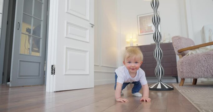 Blond toddler learning to crawl and stand on wooden floor in bright modern living room with armchair, floor lamp, cabinet, and soft rug