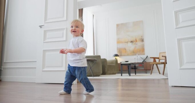 Blond toddler taking first steps across a bright modern living room with wooden floor, soft daylight, white walls, and cozy home decor