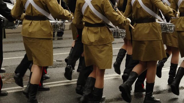 Uniformed drummers in a coordinated military-style parade march across a wet city street, showcasing synchronized steps, polished boots, and snare drums in disciplined procession