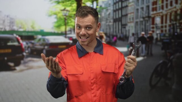 Man handyman holding wrench, pointing finger and gesturing with right hand on street near parked cars and bicycles; confidence reliability.