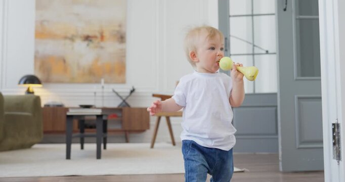 Curious toddler learning to walk across a bright modern living room while holding a yellow toy as mother watches and encourages nearby