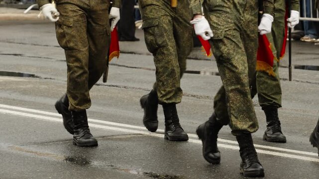 Close-up of soldiers marching in formation during a military parade on a wet city street, showcasing polished boots, mixed camouflage and dress uniforms, synchronized steps, and crowd barricades.
