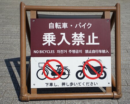 A ground-level sign prohibiting bicycles and motorcycles, instructing users to dismount and walk, displayed on a paved surface