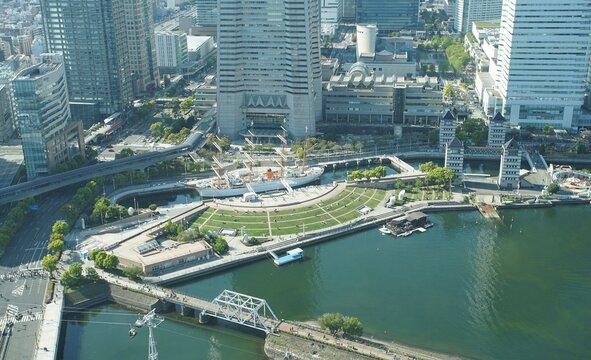 An aerial view of the Minato Mirai district in Yokohama, featuring a docked sailing ship, a grassy park, modern buildings, and a bridge over water